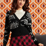 A woman wearing a black cardigan with white skeleton motifs over a white ribbed top and a red and black plaid skirt, posed against a retro orange and brown patterned wallpaper background.