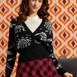 A woman wearing a black cardigan with white skeleton motifs over a white ribbed top and a red and black plaid skirt, posed against a retro orange and brown patterned wallpaper background.