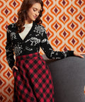 A woman posing in a black knit sweater with white skeleton motifs and a red and black buffalo plaid skirt, set against a retro orange and white patterned wall.