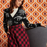 A woman posing in a black knit sweater with white skeleton motifs and a red and black buffalo plaid skirt, set against a retro orange and white patterned wall.
