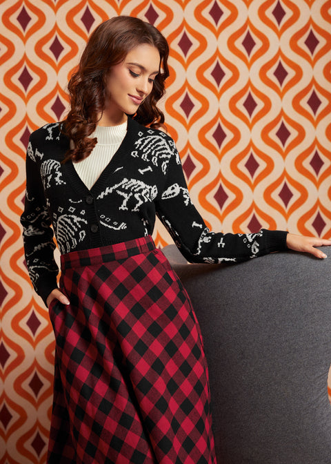 A woman posing in a black knit sweater with white skeleton motifs and a red and black buffalo plaid skirt, set against a retro orange and white patterned wall.