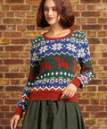 A woman wearing a festive Christmas sweater with a blue, green, and red Fair Isle pattern featuring snowflakes and dinosaurs, paired with an olive green skirt, standing against a brick wall.
