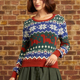 A woman wearing a festive Christmas sweater with a blue, green, and red Fair Isle pattern featuring snowflakes and dinosaurs, paired with an olive green skirt, standing against a brick wall.