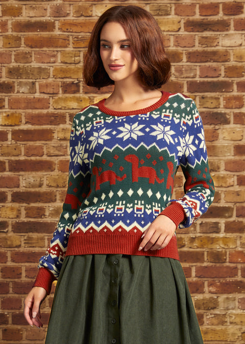 A woman wearing a festive Christmas sweater with a blue, green, and red Fair Isle pattern featuring snowflakes and dinosaurs, paired with an olive green skirt, standing against a brick wall.