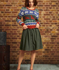 A woman stands in front of a brick wall wearing a festive multicolor Christmas sweater with a green button-front midi skirt and red Mary Jane shoes.