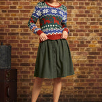 A woman stands in front of a brick wall wearing a festive multicolor Christmas sweater with a green button-front midi skirt and red Mary Jane shoes.