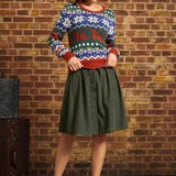 A woman stands in front of a brick wall wearing a festive multicolor Christmas sweater with a green button-front midi skirt and red Mary Jane shoes.