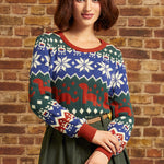 A woman wearing a festive Christmas sweater with a blue, green, and red holiday pattern, standing against a rustic brick wall.