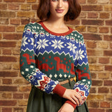 A woman wearing a festive Christmas sweater with a blue, green, and red holiday pattern, standing against a rustic brick wall.
