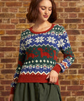 A woman wearing a festive Christmas sweater with red, green, and blue colors, featuring snowflakes and dinosaurs, standing against a brick wall.