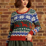 A woman wearing a festive Christmas sweater with red, green, and blue colors, featuring snowflakes and dinosaurs, standing against a brick wall.