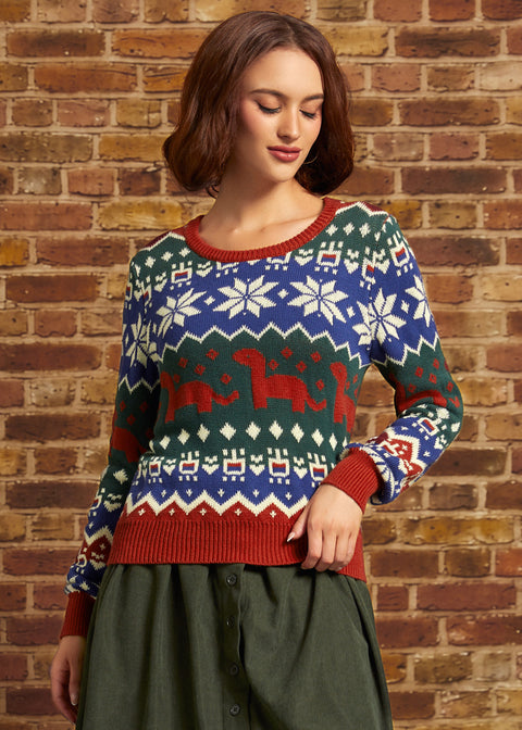 A woman wearing a festive Christmas sweater with red, green, and blue colors, featuring snowflakes and dinosaurs, standing against a brick wall.