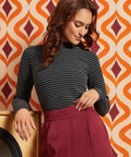 A woman wearing a dark gray and black striped mock-neck long-sleeve top and a burgundy pleated skirt, posing in front of a retro orange, beige, and burgundy patterned wallpaper background.