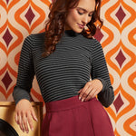 A woman wearing a dark gray and black striped mock-neck long-sleeve top and a burgundy pleated skirt, posing in front of a retro orange, beige, and burgundy patterned wallpaper background.