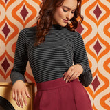 A woman wearing a dark gray and black striped mock-neck long-sleeve top and a burgundy pleated skirt, posing in front of a retro orange, beige, and burgundy patterned wallpaper background.