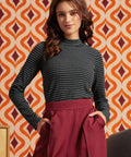 A woman modeling a dark gray and black striped mock neck long-sleeve top paired with a high-waisted burgundy skirt, standing in front of a retro orange and cream patterned wallpaper background.