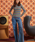 A woman models a retro 1970s-style outfit featuring a multicolor horizontally striped mock-neck sweater and wide-leg blue jeans with embroidered stripes