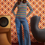 A woman models a retro 1970s-style outfit featuring a multicolor horizontally striped mock-neck sweater and wide-leg blue jeans with embroidered stripes