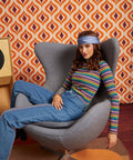 A woman wearing a colorful striped long-sleeve top, blue jeans, and a blue headband sits in a gray egg-shaped armchair in front of a retro orange and brown patterned wallpaper background.