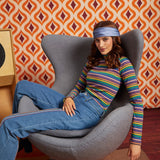 A woman wearing a colorful striped long-sleeve top, blue jeans, and a blue headband sits in a gray egg-shaped armchair in front of a retro orange and brown patterned wallpaper background.