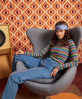 A woman wearing a colorful striped long-sleeve mock neck top, blue jeans, and a blue headband sits in a gray lounge chair against a retro orange and brown patterned wallpaper background.