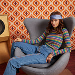 A woman wearing a colorful striped long-sleeve mock neck top, blue jeans, and a blue headband sits in a gray lounge chair against a retro orange and brown patterned wallpaper background.