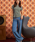 A woman modeling a retro-themed outfit featuring a green and orange striped t-shirt, blue wide-leg jeans with a distinctive curved seam detail, and a colorful patterned headband, posed in front of a retro orange and brown patterned wallpaper background.