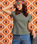 A woman wearing a short-sleeve mock neck top with thin multicolor horizontal stripes (green, orange, blue, red) and blue jeans, posing in front of a retro orange and brown patterned wallpaper background.