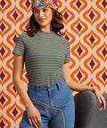 A woman wearing a short-sleeve ribbed knit top with thin multicolor horizontal stripes, paired with blue jeans and a colorful patterned headband, posed in front of a retro orange and brown patterned wall.