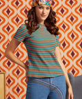 A woman wearing a short-sleeve crewneck t-shirt with horizontal stripes in teal, orange, yellow, and green, paired with blue jeans and a colorful patterned headband, posed against a retro orange and cream patterned wallpaper background.