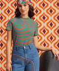 A woman wearing a short-sleeve striped knit top with blue jeans and a colorful patterned headband, posed against a retro orange, cream, and burgundy geometric patterned wall.