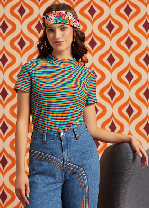 A woman wearing a short-sleeve striped knit top with blue jeans and a colorful patterned headband, posed against a retro orange, cream, and burgundy geometric patterned wall.