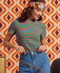 A woman wearing a short-sleeve ribbed mock neck top with thin multicolor horizontal stripes (green, orange, red, blue) and blue jeans, paired with a colorful floral headband, standing in front of a retro orange and brown patterned wallpaper background.