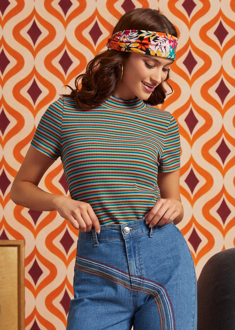 A woman wearing a short-sleeve ribbed mock neck top with thin multicolor horizontal stripes (green, orange, red, blue) and blue jeans, paired with a colorful floral headband, standing in front of a retro orange and brown patterned wallpaper background.