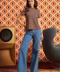 A woman stands in a retro-styled room wearing a ModCloth brown and orange striped short-sleeve t-shirt and blue wide-leg jeans with a distinctive rainbow stripe detail down the side.
