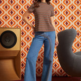 A woman stands in a retro-styled room wearing a ModCloth brown and orange striped short-sleeve t-shirt and blue wide-leg jeans with a distinctive rainbow stripe detail down the side.