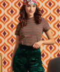 A woman modeling a short-sleeve mockneck striped t-shirt in brown, beige, and orange tones, paired with dark green floral velvet pants and a colorful tropical print headband, set against a retro orange and cream patterned wallpaper background.