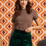 A woman modeling a short-sleeve mockneck striped t-shirt in brown, beige, and orange tones, paired with dark green floral velvet pants and a colorful tropical print headband, set against a retro orange and cream patterned wallpaper background.