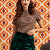 A woman modeling a short-sleeve mockneck striped t-shirt in brown, beige, and orange tones, paired with dark green floral velvet pants and a colorful tropical print headband, set against a retro orange and cream patterned wallpaper background.