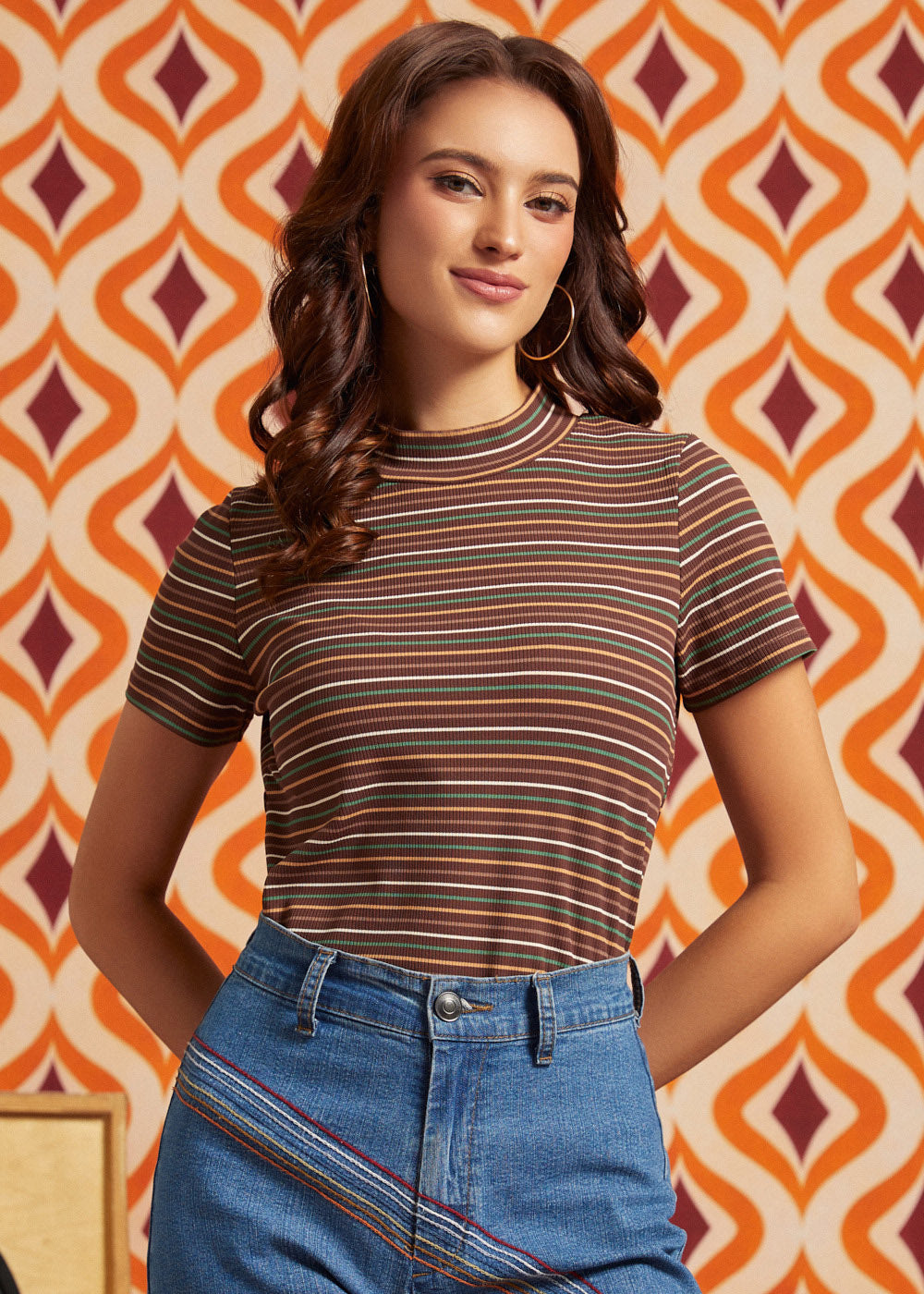 A woman wearing a short-sleeve mock neck t-shirt with thin horizontal stripes in brown, orange, and white, paired with blue jeans, standing in front of a retro orange and cream patterned wallpaper background.