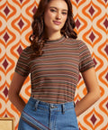 A woman wearing a short-sleeve mock neck t-shirt with thin horizontal stripes in brown, orange, and white, paired with blue jeans, standing in front of a retro orange and cream patterned wallpaper background.