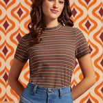 A woman wearing a short-sleeve mock neck t-shirt with thin horizontal stripes in brown, orange, and white, paired with blue jeans, standing in front of a retro orange and cream patterned wallpaper background.