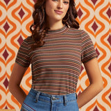 A woman wearing a short-sleeve mock neck t-shirt with thin horizontal stripes in brown, orange, and white, paired with blue jeans, standing in front of a retro orange and cream patterned wallpaper background.