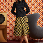 A woman models a black long-sleeve lace blouse paired with a yellow and black checkered midi skirt, standing in a room with a retro orange and brown patterned wall, a gray chair, and a wooden speaker.