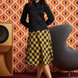 A woman models a black long-sleeve lace blouse paired with a yellow and black checkered midi skirt, standing in a room with a retro orange and brown patterned wall, a gray chair, and a wooden speaker.