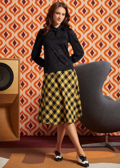 A woman models a black long-sleeve lace blouse paired with a yellow and black checkered midi skirt, standing in a room with a retro orange and brown patterned wall, a gray chair, and a wooden speaker.