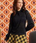 Woman wearing a black floral lace tie-neck top with long puff sleeves and a yellow and black check midi skirt.