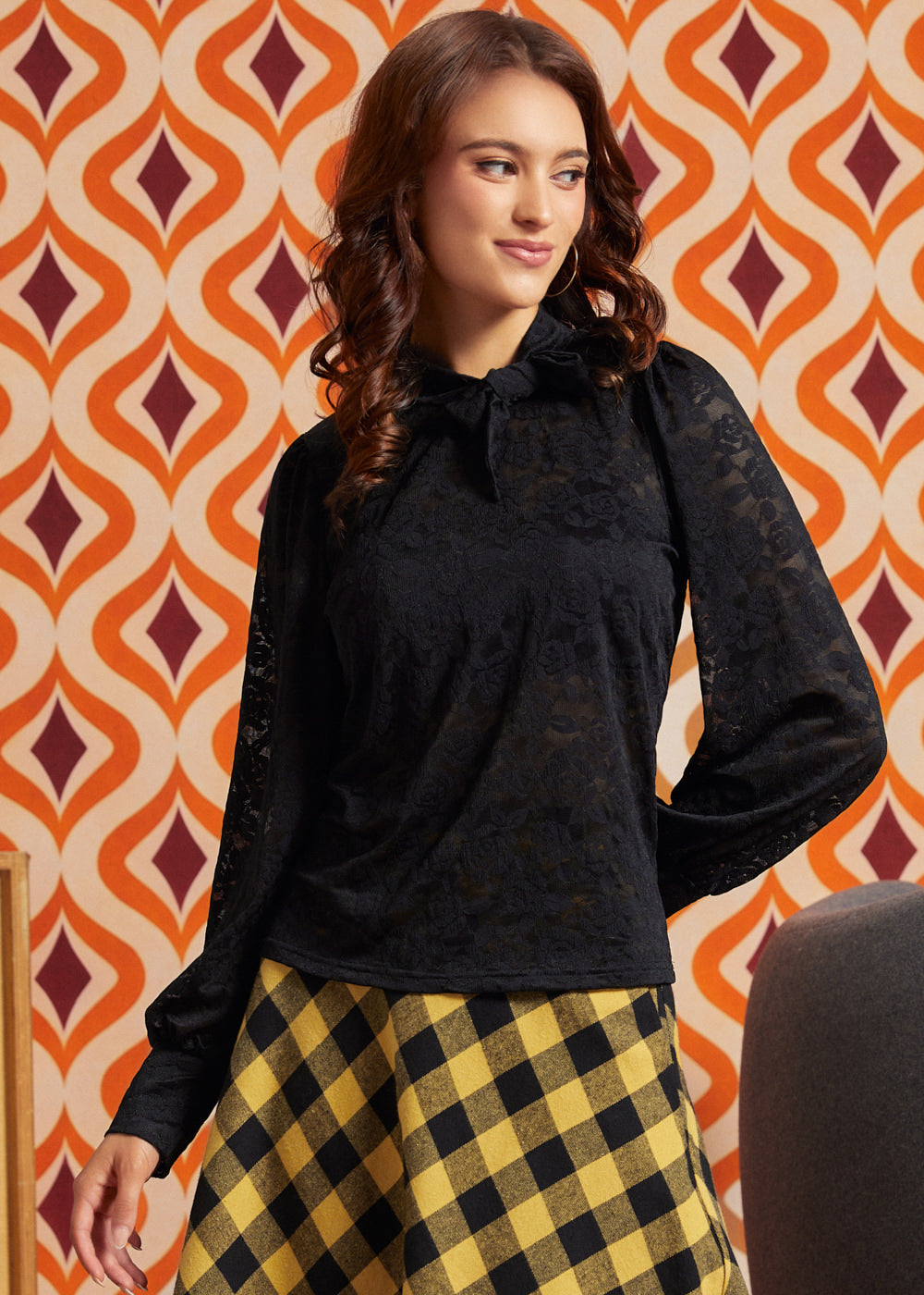 Woman wearing a black floral lace tie-neck top with long puff sleeves and a yellow and black check midi skirt.