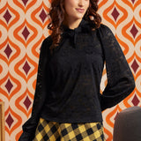 Woman wearing a black floral lace tie-neck top with long puff sleeves and a yellow and black check midi skirt.