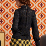 A woman shown from the back wearing a black lace long-sleeve blouse and a yellow and black checkered skirt, standing in front of a retro orange and cream patterned wallpaper.